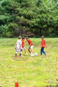 Four children, three in orange shirts and one in gray, playing soccer on a grassy field.