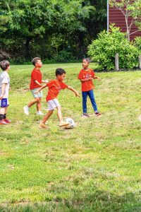 Four children playing soccer on a grassy field near bushes and a red building.