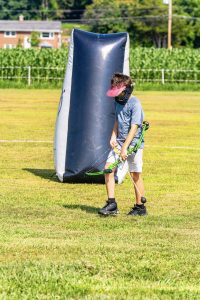 Child wearing protective mask and visor holding a green bow near an inflatable bunker on grass
