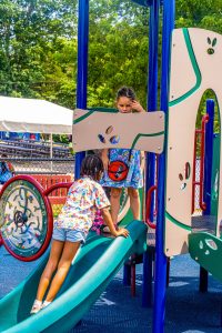 Two children playing on colorful playground slide with trees and tents in the background