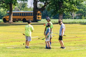 Four people wearing face shields holding bows on a grassy field near a yellow school bus.