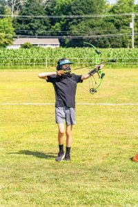 Person wearing protective mask aiming a green compound bow with an arrow on a grassy field