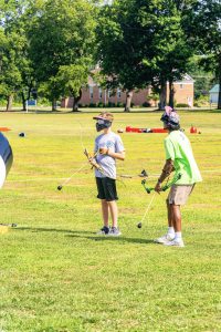 Two people wearing masks holding bows with foam-tipped arrows on a grassy field.