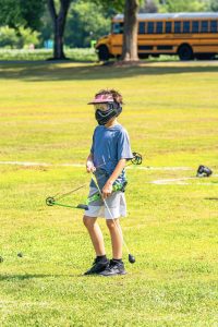 Boy in protective mask and visor holding green compound bow on grass near school bus