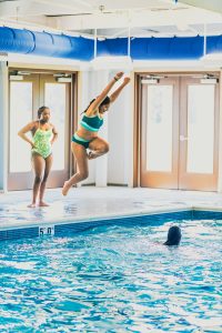 Three girls at an indoor pool; one jumping in, one standing, one swimming, large doors visible.
