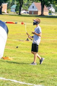 Young person wearing a protective mask holding a bow and arrow on a grassy field outdoors.