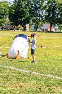 Boy in protective mask holding paintball gun near inflatable bunker on grassy field.