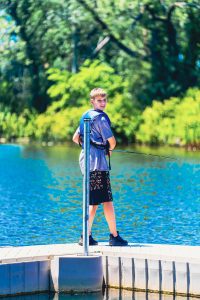 Boy wearing a blue life vest fishing from a dock on a lake with green trees in the background