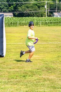 Boy in yellow shirt and gray shorts running on grass holding a purple bandana and yellow ball.