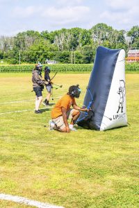 Three people wearing protective gear practicing archery near an inflatable UPSHOT ADVENTURES barrier on a field.
