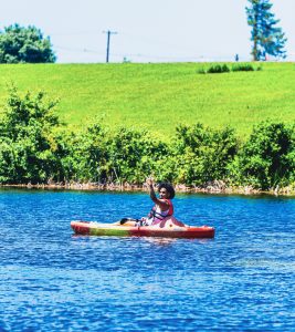 Person in a red life jacket paddling a kayak on a lake with green grass and bushes nearby