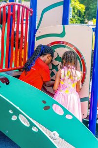 Two children with braided hair playing on colorful playground equipment outdoors.