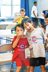 Two boys in day camp shirts pose indoors, one holding a snack bag, others eating popsicles.