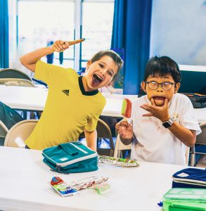 Two children sitting at a table indoors, making playful faces while holding popsicles.
