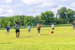 Children playing kickball on a grassy field with volleyball nets and trees in the background.