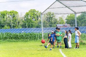 Children and an adult near a baseball backstop with solar panels and cornfield in background