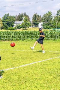 Person in black sportswear kicking a red ball on grassy field with cornfields and houses.