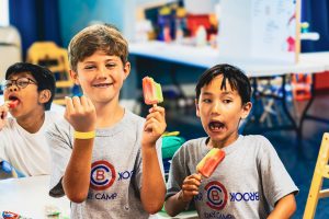 Three boys indoors eating colorful popsicles, two wearing matching Brook Day Camp shirts.