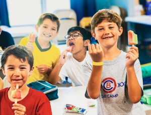 Four boys indoors smiling and eating colorful popsicles at a table.