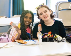 Two girls sitting at a table eating popsicles with pizza, popcorn, and a juice pouch.