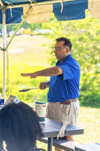 Man in blue shirt holding spatula with food stands under canopy beside table with items and skewers.