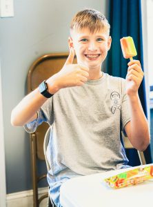 Boy with braces in gray t-shirt holding a multi-colored popsicle and giving thumbs up at table.