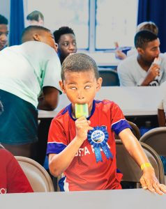 Boy in red shirt with birthday ribbon eating a yellow and green popsicle at a table.