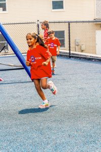 Three children in red camp t-shirts playing on blue playground surface with chain-link fence.