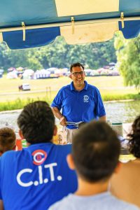 Man in blue shirt holding tongs under a striped canopy speaking to seated children outdoors.