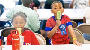 Two children wearing red shirts are sitting at a table eating green popsicles indoors.