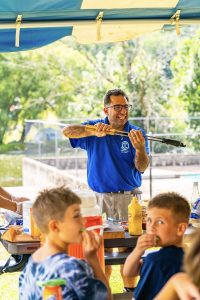 Man in blue shirt grilling food with children eating at picnic table outdoors under canopy.