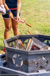 Two people roasting hot dogs and slices of meat over a metal fire pit with star cutouts.