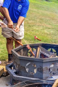 Person roasting hot dogs and bologna on skewers over a star-cutout metal fire pit outdoors.