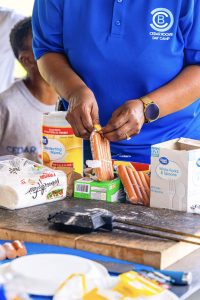 Person in a blue Cedar Brook Day Camp shirt holding hot dogs on a table with utensils and wipes.