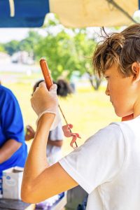 Boy in white t-shirt wearing a glove holding a sausage and raw meat on a skewer outdoors.