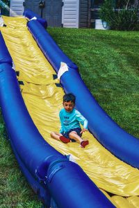 Young boy in blue shirt and red shoes sliding down a yellow and blue inflatable slip-and-slide.