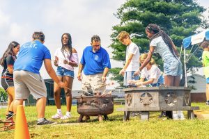 People around fire pits roasting food on sticks outdoors in a grassy area with trees and tents.