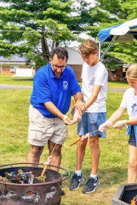 A man and two children roasting hot dogs over a fire pit outdoors on grass.