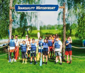Group of people in life jackets holding paddles standing under Tranquility Waterfront sign on grass.