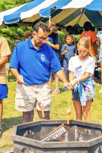 Adult and child roasting hot dogs over fire pit at outdoor event with people under tent.