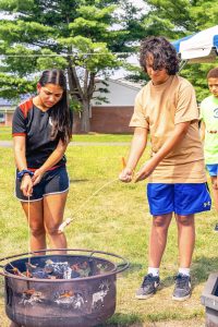 Teenagers roasting hot dogs over a fire pit outdoors with grass, trees, and buildings behind.