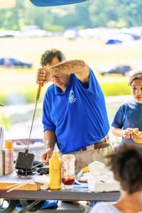 Man in blue Cedar Woods Day Camp shirt making sandwiches outdoors with children nearby.