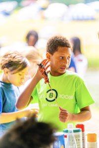 Boy in bright green day camp shirt holds grilled skewer near table with bottles and other children.