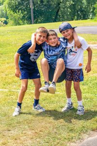 Three boys outdoors on grass, two standing and holding the third boy off the ground by his arms.