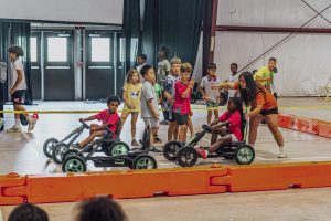 Children indoors with pedal go-karts, an adult gives instructions near orange barriers.