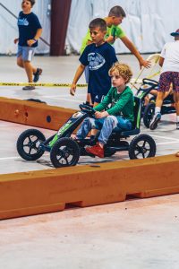 Children riding and walking around pedal go-karts on an indoor track with orange barriers.