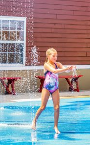 Girl in horse swimsuit playing in water jets at splash pad with red building background