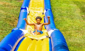 Boy in blue swim trunks sliding down a yellow and blue inflatable water slide on grass.