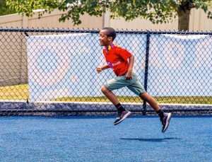 Boy wearing red shirt, green shorts, and flower lei running on a blue track by a fence.