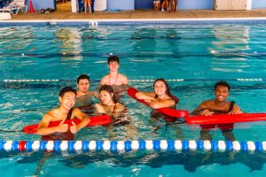 Six young lifeguards in a pool holding red rescue tubes near lane dividers.